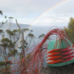 A chevron-patterned telephone wire basket on a stump of wood, with a view of Maria Island in the distance and a rainbow overhead.