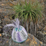 Purple and white chevron patterned wire basket in progress, on a small boulder with a Pandanus plant behind it.