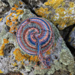 Blue, orange, white and black telephone wire basket in progress on a rock richly covered in lichens.